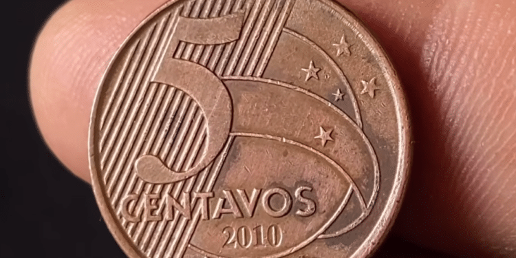 Close-up detalhado do reverso de uma moeda de 5 centavos de real do ano de 2010, sendo segurada pelos dedos de uma pessoa contra um fundo escuro.