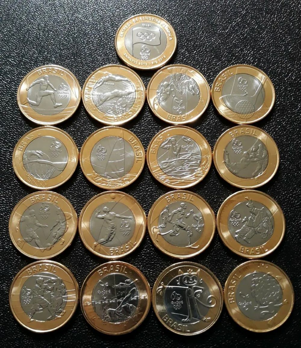 Dezessete moedas de 1 Real comemorativas dos Jogos Rio 2016, organizadas em uma pirâmide invertida no topo de quatro colunas de quatro moedas cada. No topo da pirâmide, a moeda da 'Bandeira Olímpica'. As outras exibem diversas modalidades esportivas e os mascotes Tom e Vinícius.