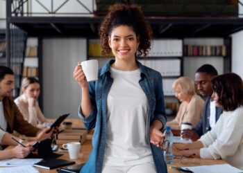 Jovens participando de uma reunião em ambiente de escritório, com foco no trabalho colaborativo, enquanto alguns estão com xícaras e outros com materiais de escritório.