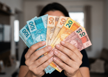 Pessoa segura várias notas de real abertas em leque com ambiente de sala ao fundo