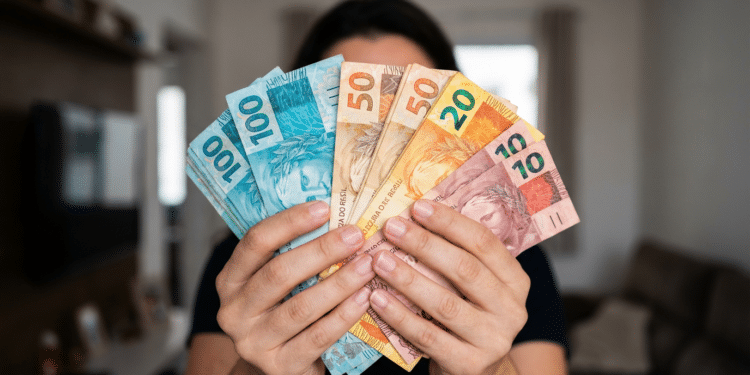 Pessoa segura várias notas de real abertas em leque com ambiente de sala ao fundo