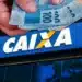 Pessoa segura várias cédulas de reais sobre a fachada da Caixa Econômica Federal