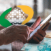 Mãos de idoso usando celular ao lado de notas de real e logomarca do INSS