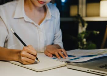 Jovem escrevendo em caderno à noite com documentos e notebook representando como estruturar a redação do Enem 2026