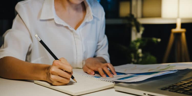 Jovem escrevendo em caderno à noite com documentos e notebook representando como estruturar a redação do Enem 2026
