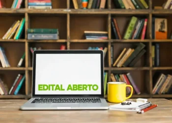 Computador com a tela mostrando a frase 'EDITAL ABERTO', xícara amarela ao lado e livros ao fundo — Prefeitura anuncia 100 vagas com salários de até R$ 4 mil.