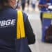 Mulher com colete do IBGE segura bolsa amarela enquanto aplica pesquisa em área urbana