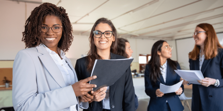 Cinco mulheres com ternos sociais trocando documentos e sorrindo em escritório