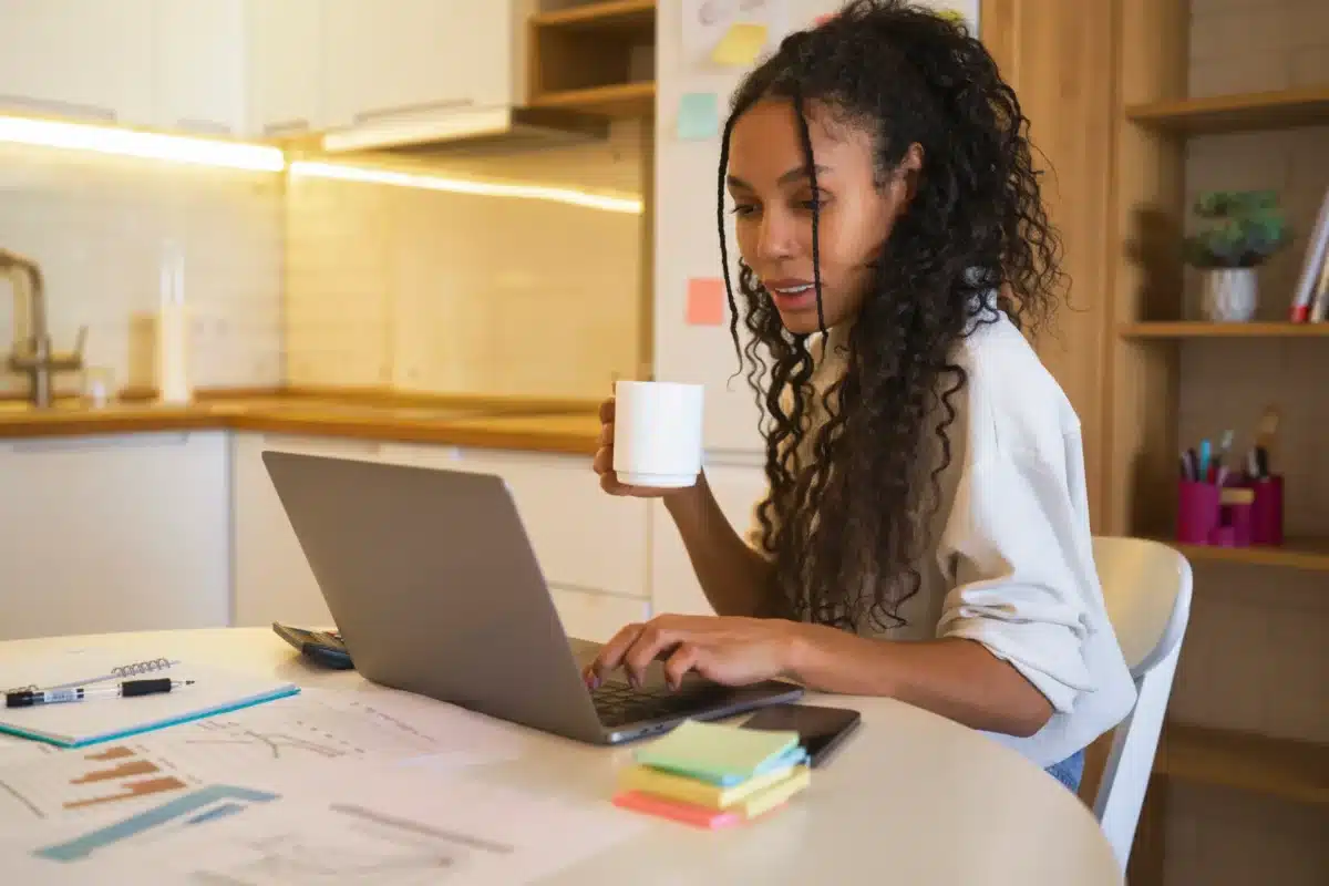 Mulher trabalhando em home office com notebook e xícara de café, em um ambiente doméstico organizado.