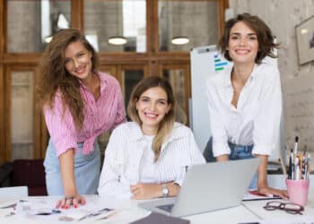 Três mulheres jovens sorrindo apoiadas sobre mesa de trabalho com notebook, materiais criativos e pincéis em escritório iluminado