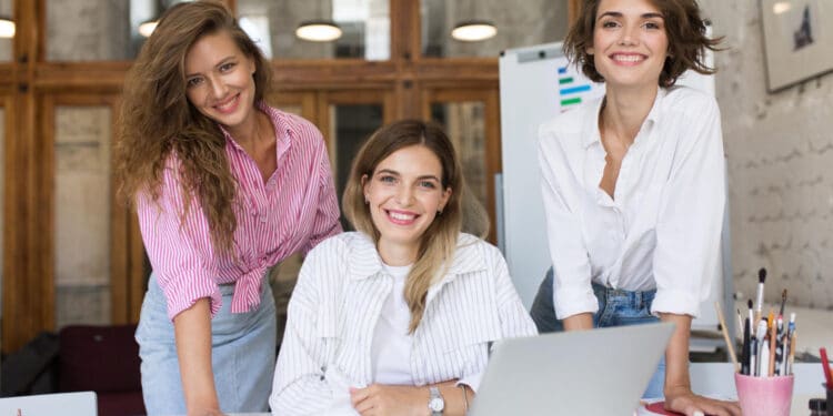 Três mulheres jovens sorrindo apoiadas sobre mesa de trabalho com notebook, materiais criativos e pincéis em escritório iluminado