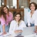 Três mulheres jovens sorrindo apoiadas sobre mesa de trabalho com notebook, materiais criativos e pincéis em escritório iluminado