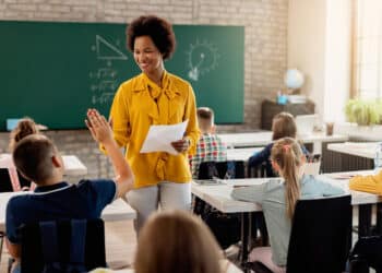 Professora sorrindo em sala de aula com alunos levantando a mão — proposta de novos adicionais salariais para professores 2026