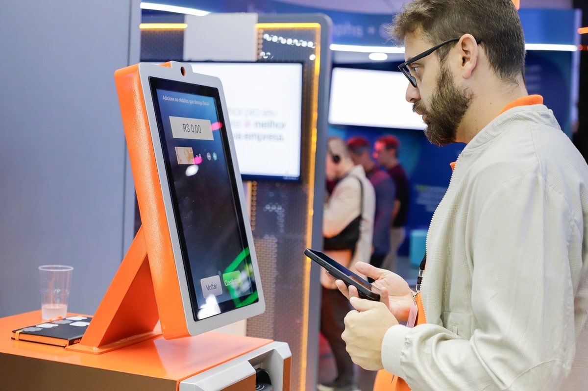 Homem usando smartphone em frente a terminal digital laranja do Banco Inter para acessar as condições especiais de renegociação de dívidas com 98% de desconto