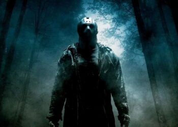 Figura sombria do personagem Jason Voorhees, de Sexta-Feira 13, usando sua icônica máscara de hóquei. Ele está em pé em uma floresta nebulosa e escura à noite, iluminada por uma luz azulada vinda de trás das árvores.