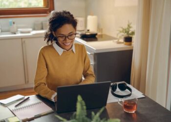 Mulher sorrindo enquanto trabalha em home office, com notebook, chá e fones de ouvido em um ambiente doméstico iluminado
