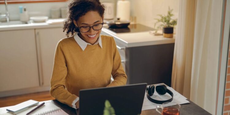 Mulher sorrindo enquanto trabalha em home office, com notebook, chá e fones de ouvido em um ambiente doméstico iluminado