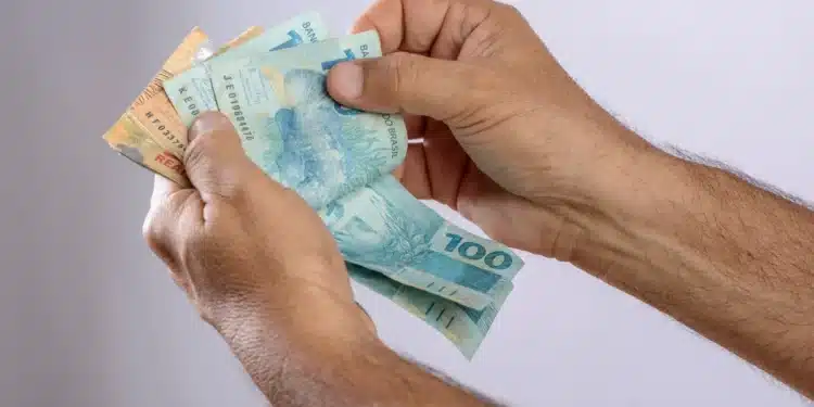 Pessoa segurando notas de reais, incluindo valores de R$ 100, R$ 50 e R$ 20, com foco em dinheiro em notas brasileiras.