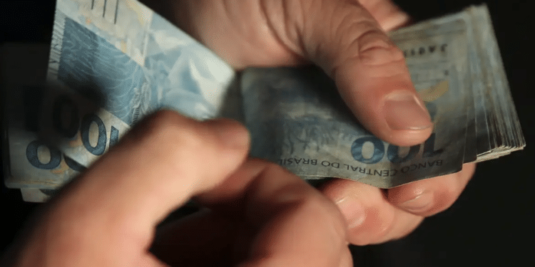 Mãos contando cédulas de R$ 100 sobre fundo escuro representando o Salário mínimo 2027