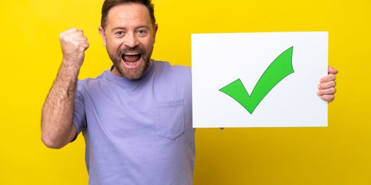 Homem celebra vitória segurando placa com sinal de aprovação verde.