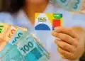 Cartão do Bolsa Família sendo segurado por uma pessoa, com cédulas de dinheiro de R$ 100, R$ 50 e R$ 20 ao fundo, representando os pagamentos do programa em abril de 2026.