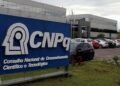 Logotipo do CNPq, localizado na sede do Conselho Nacional de Desenvolvimento Científico e Tecnológico, com fundo de carros estacionados.