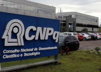 Logotipo do CNPq, localizado na sede do Conselho Nacional de Desenvolvimento Científico e Tecnológico, com fundo de carros estacionados.
