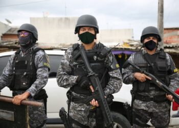Três policiais militares do Amazonas, em uniforme tático e armados, estão em uma operação de segurança, com um cenário urbano ao fundo.