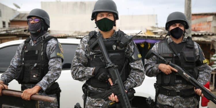 Três policiais militares do Amazonas, em uniforme tático e armados, estão em uma operação de segurança, com um cenário urbano ao fundo.