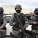 Três policiais militares do Amazonas, em uniforme tático e armados, estão em uma operação de segurança, com um cenário urbano ao fundo.