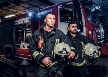 Bombeiros em frente a um caminhão de bombeiros, prontos para uma emergência, durante a noite, com equipamentos de proteção.