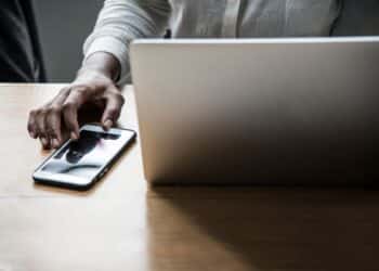 Pessoa trabalhando em um laptop com um smartphone ao lado, representando as mudanças nas regras de acesso aos serviços digitais da Receita Federal, incluindo autenticação via conta gov.br e representação digital.