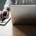 Pessoa trabalhando em um laptop com um smartphone ao lado, representando as mudanças nas regras de acesso aos serviços digitais da Receita Federal, incluindo autenticação via conta gov.br e representação digital.