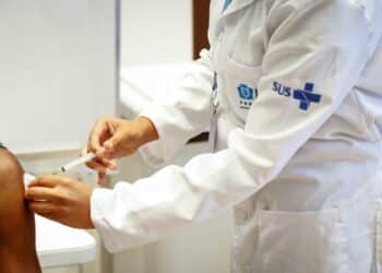 Profissional de saúde aplicando vacina em um paciente, com uniforme do SUS, durante a Semana de Vacinação nas Américas.