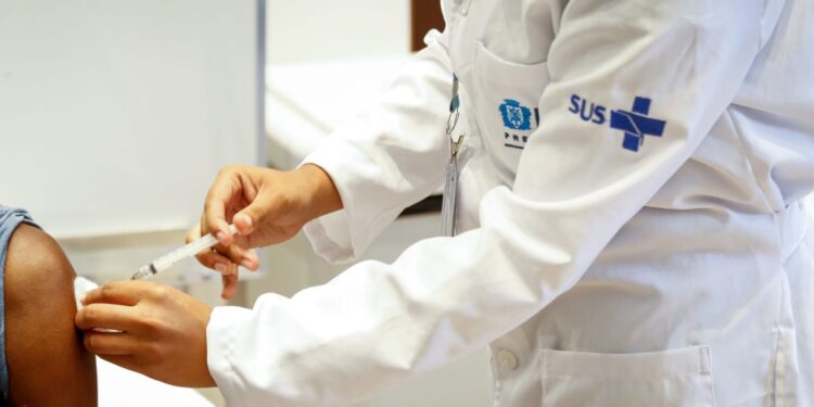 Profissional de saúde aplicando vacina em um paciente, com uniforme do SUS, durante a Semana de Vacinação nas Américas.