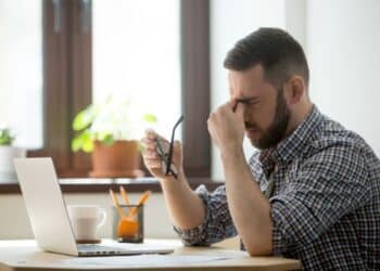 Profissional sentado à mesa de trabalho com laptop, segurando óculos e pressionando os olhos, demonstrando cansaço e sobrecarga.