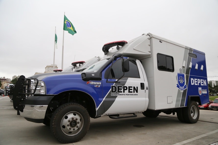 Veículo blindado com identificação do DEPEN Paraná estacionado ao lado das bandeiras do Brasil e do estado