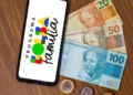 Celular exibindo logotipo do Programa Bolsa Família ao lado de notas de dinheiro representando antecipação do pagamento em abril