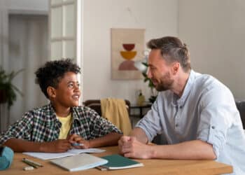 Pai e filho conversam sorridentes durante estudo em casa, com cadernos e estojo sobre a mesa