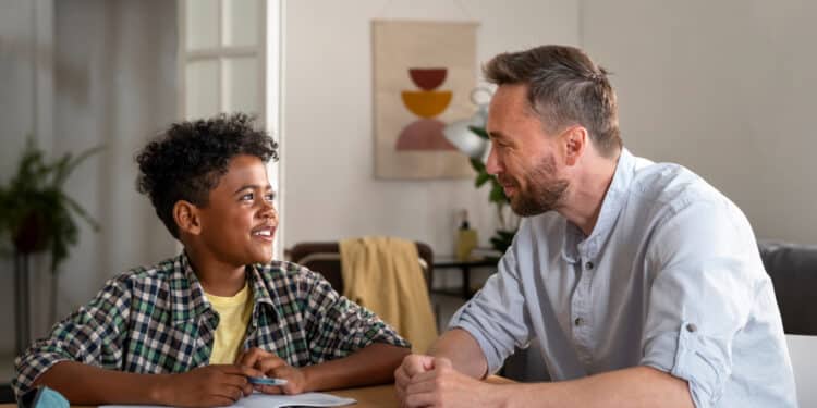 Pai e filho conversam sorridentes durante estudo em casa, com cadernos e estojo sobre a mesa