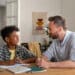 Pai e filho conversam sorridentes durante estudo em casa, com cadernos e estojo sobre a mesa
