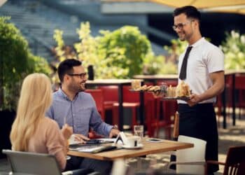 Garçom servindo bebidas e petiscos a clientes em um restaurante ao ar livre. Profissional de atendimento trabalhando com escalas 6x1.