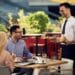 Garçom servindo bebidas e petiscos a clientes em um restaurante ao ar livre. Profissional de atendimento trabalhando com escalas 6x1.