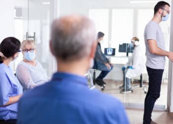 Paciente idosa aguarda atendimento ao lado de profissional de saúde em corredor de unidade hospitalar, representando o direito à prioridade para pessoas com mais de 60 anos.