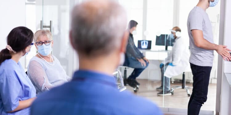 Paciente idosa aguarda atendimento ao lado de profissional de saúde em corredor de unidade hospitalar, representando o direito à prioridade para pessoas com mais de 60 anos.