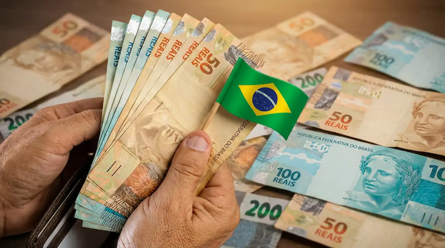 Pessoa segurando notas de real com bandeira do Brasil simbolizando economia em benefícios sociais e revisão de gastos públicos.
