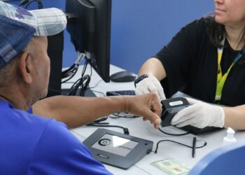 Atendente com luvas brancas realizando coleta de impressão digital de cidadão em posto de atendimento do governo