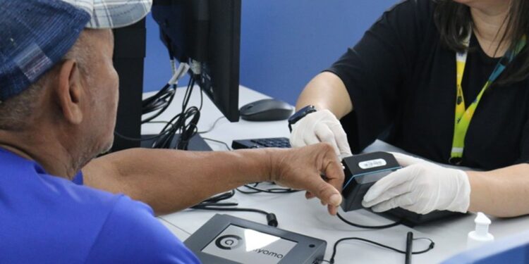 Atendente com luvas brancas realizando coleta de impressão digital de cidadão em posto de atendimento do governo