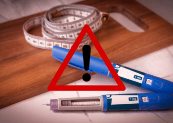 Canetas injetáveis azuis com fita métrica sobre tábua de madeira e símbolo de alerta.