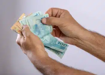 Mão segurando notas de 100 e 50 reais, representando o 13º salário e o rendimento do INSS.